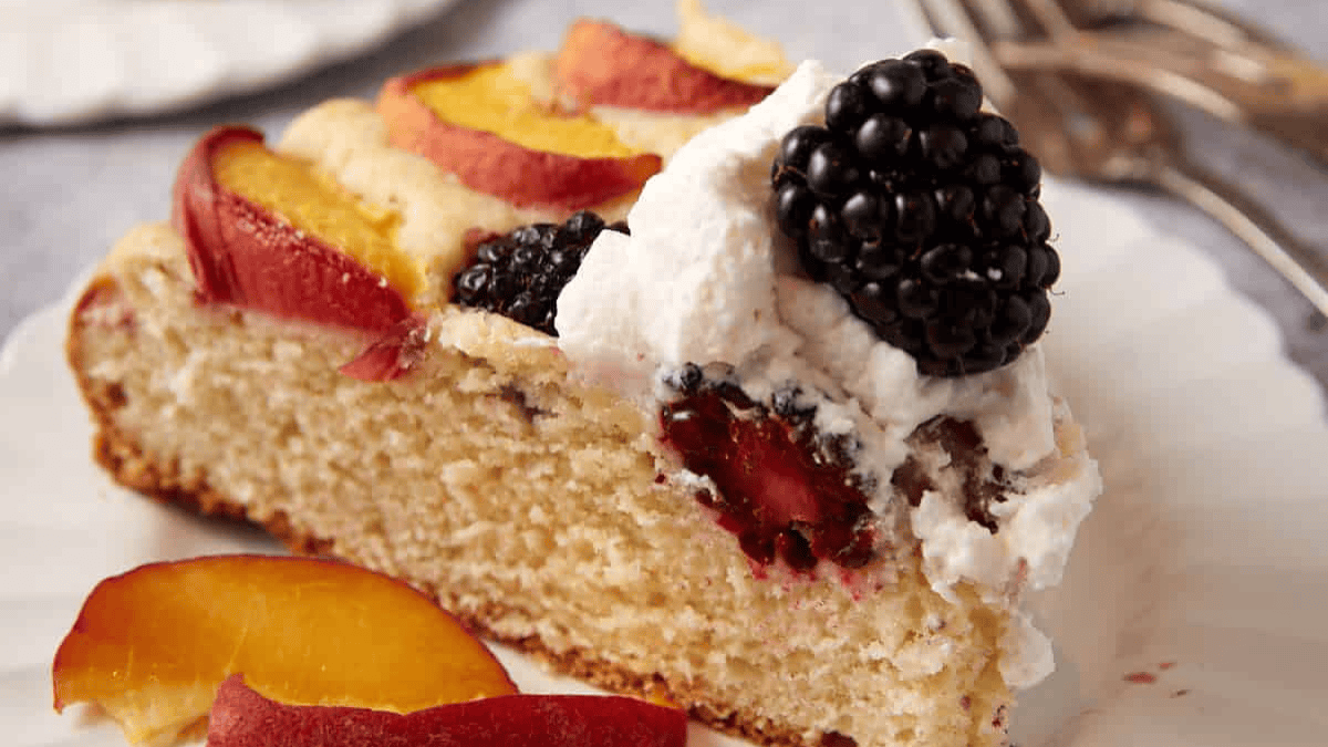 A slice of cake topped with whipped cream, fresh peach slices, and a blackberry, served on a white plate with silver forks in the background.
