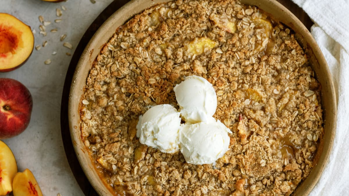 A round peach crisp topped with three scoops of vanilla ice cream sits on a light surface, surrounded by fresh peach slices and a white cloth napkin.