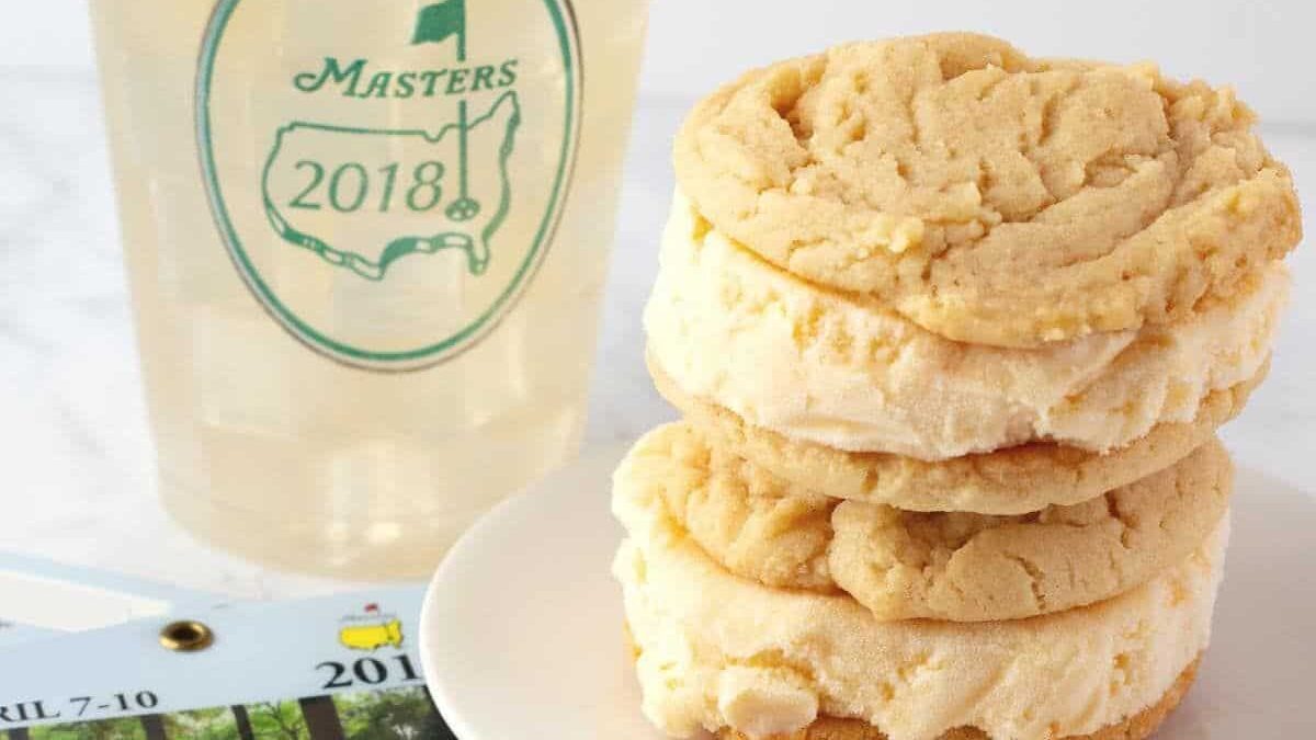 A stack of three cookies sits on a white plate next to a 2018 Masters Tournament cup filled with a yellow beverage, reminiscent of classic peach desserts. Part of a Masters badge is visible in the corner.