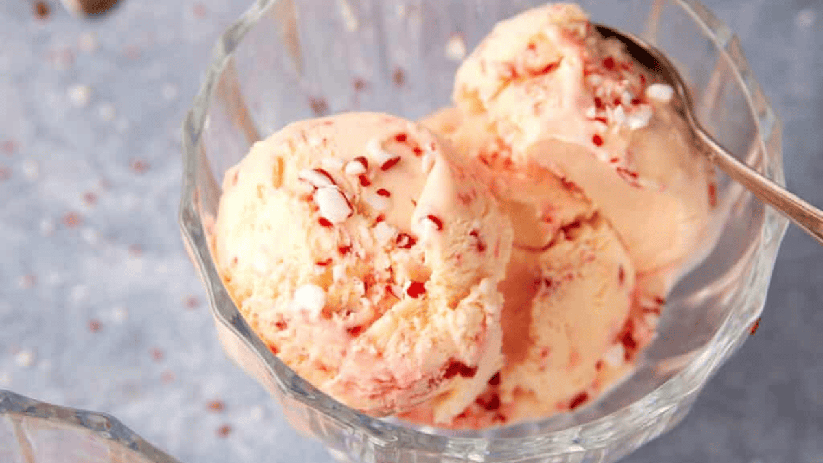Three scoops of creamy ice cream with red peppermint candy pieces in a glass bowl, with a spoon on the side; the background is light blue with white and red specks.