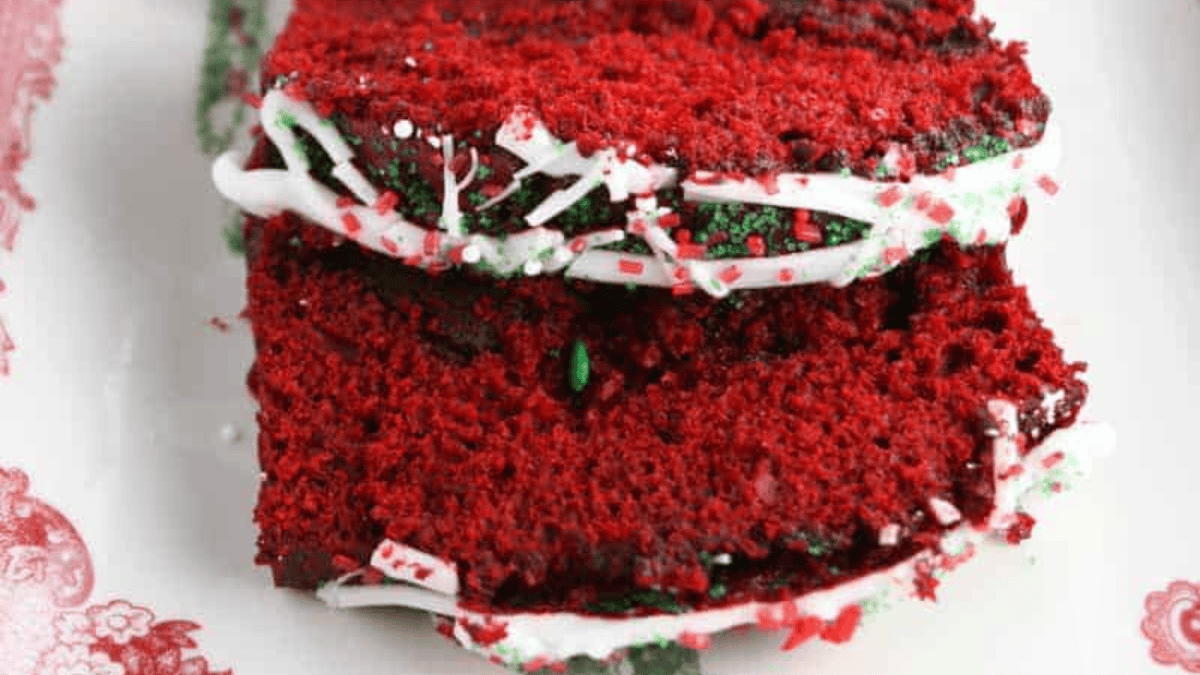 Two slices of red velvet cake topped with white icing drizzle and colorful red and green sprinkles, served on a patterned plate.
