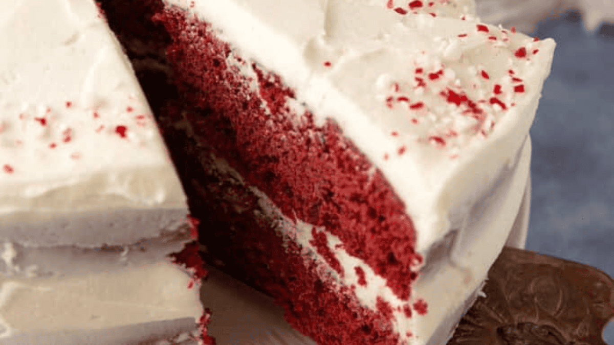 A close-up of a red velvet cake with white cream cheese frosting, topped with red sprinkles. A large slice has been cut and partially lifted from the cake, revealing its moist, layered interior.