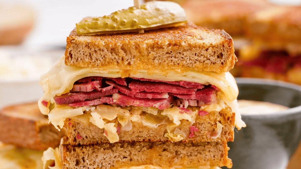 A close-up of a stacked Reuben sandwich with corned beef, melted Swiss cheese, sauerkraut, and rye bread, topped with a sliced pickle held by a toothpick.