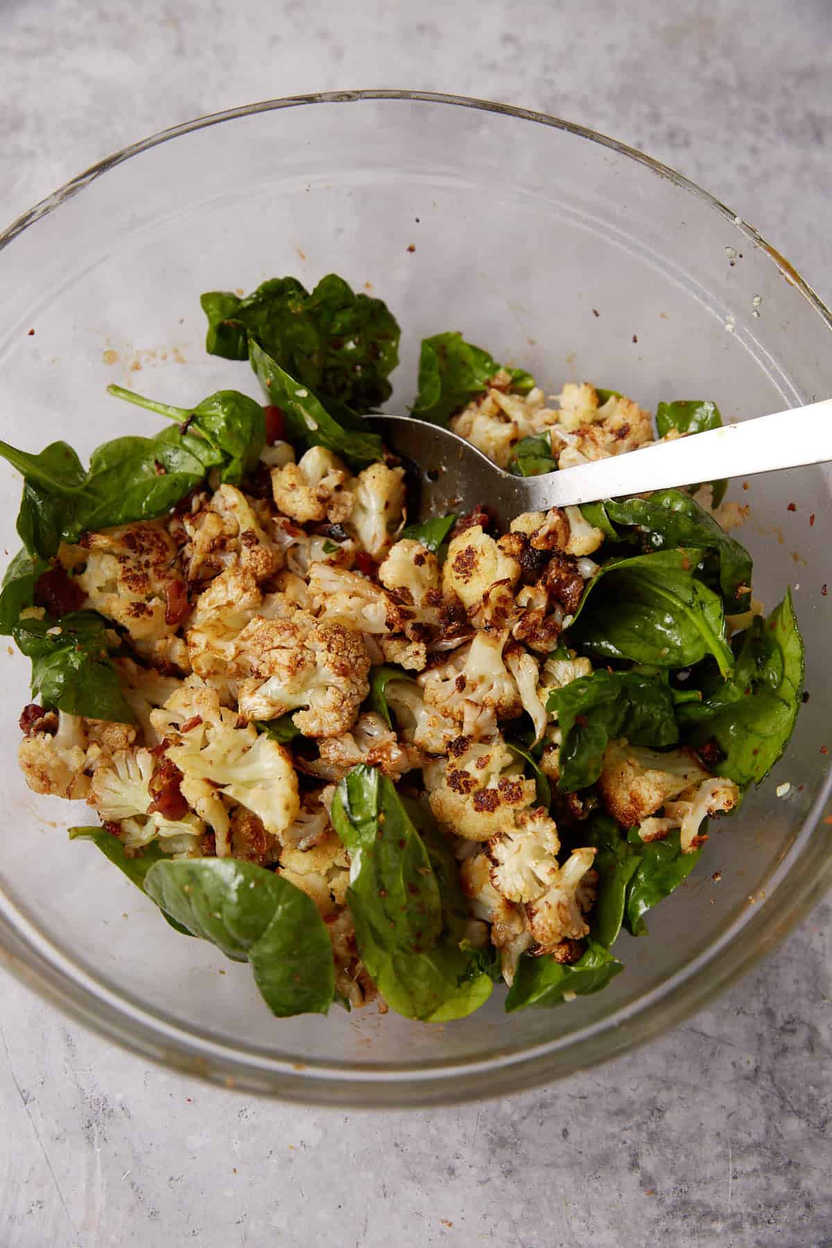A glass bowl filled with roasted cauliflower, fresh spinach leaves, and bits of what appears to be bacon, mixed together with seasoning. A metal spoon is resting in the bowl, beside a plate of savory Grape Jelly Meatballs.