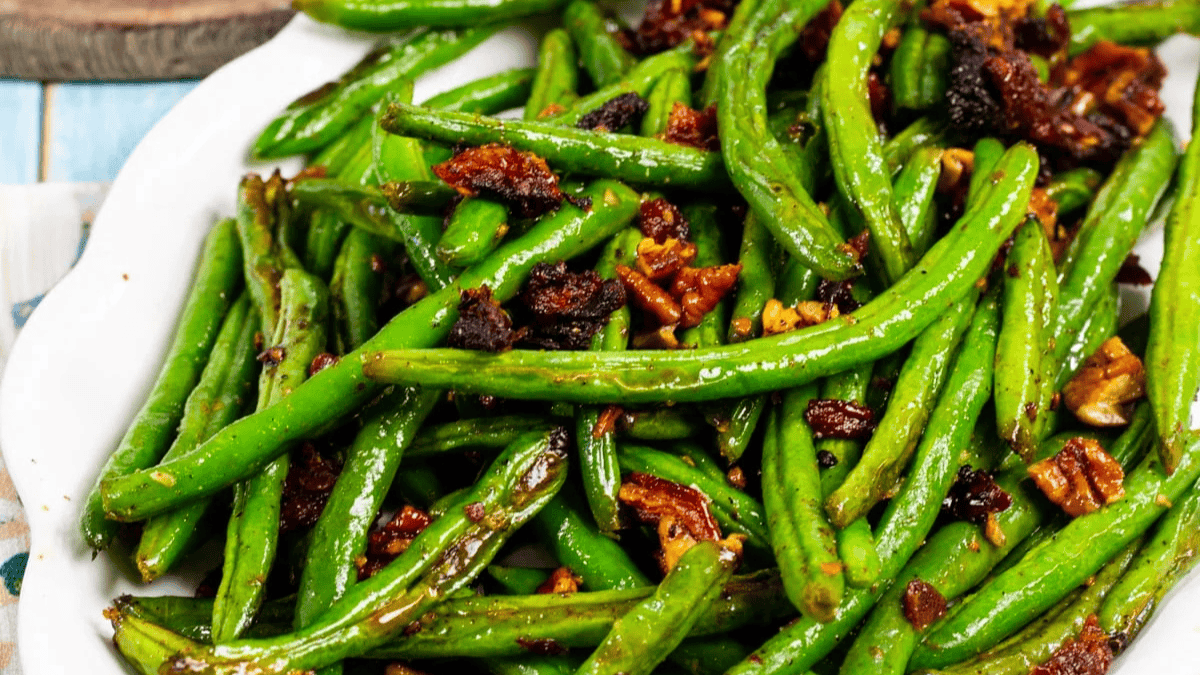 A plate of sautéed green beans mixed with crispy bacon bits and chopped pecans, served on a white dish.