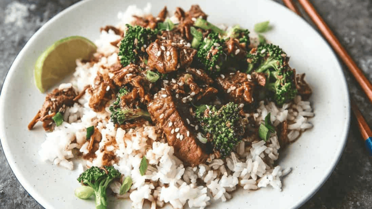 A plate of white rice topped with beef and broccoli in a savory sauce, garnished with sesame seeds and green onions, with a lime wedge on the side and chopsticks nearby.