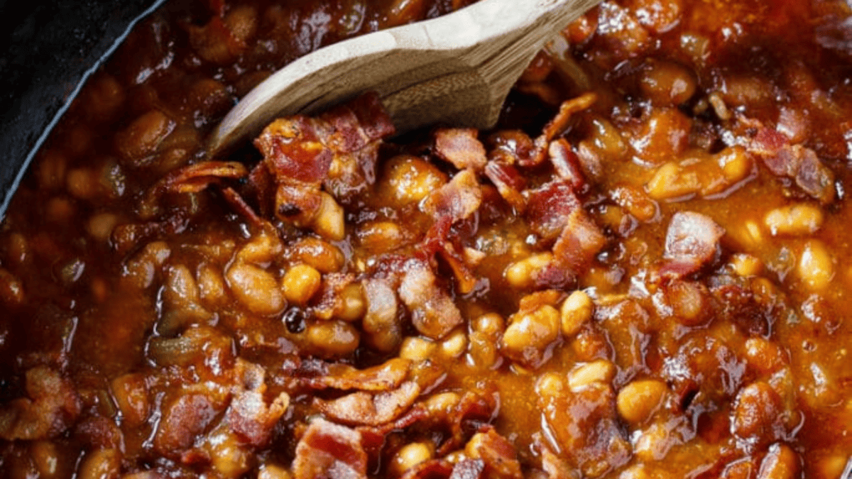 Close-up of baked beans in a rich sauce, topped with pieces of crispy bacon, being stirred with a wooden spoon.