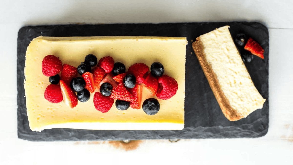 A rectangular cheesecake topped with fresh raspberries, strawberries, and blueberries sits on a black slate board, with a single sliced piece placed next to it.