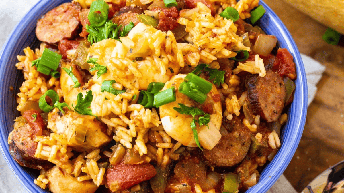 A bowl of jambalaya featuring rice, sausage, chicken, tomatoes, peppers, and onions, garnished with chopped green onions and parsley.