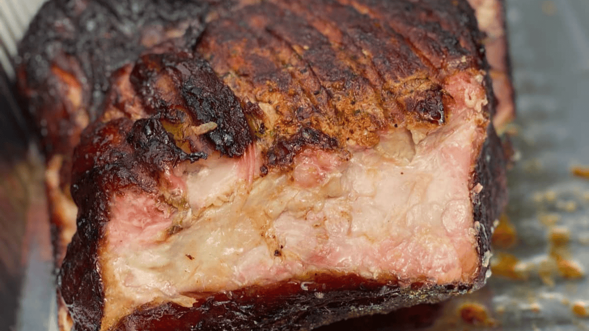 A close-up of a smoked, charred piece of meat with a browned, crispy crust and juicy, pinkish layers of fat and muscle, resting on a metal tray.