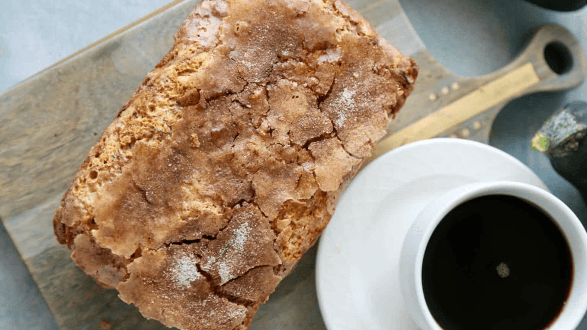 A loaf of bread with a crackly, sugared crust sits on a wooden cutting board next to a white cup filled with black coffee.