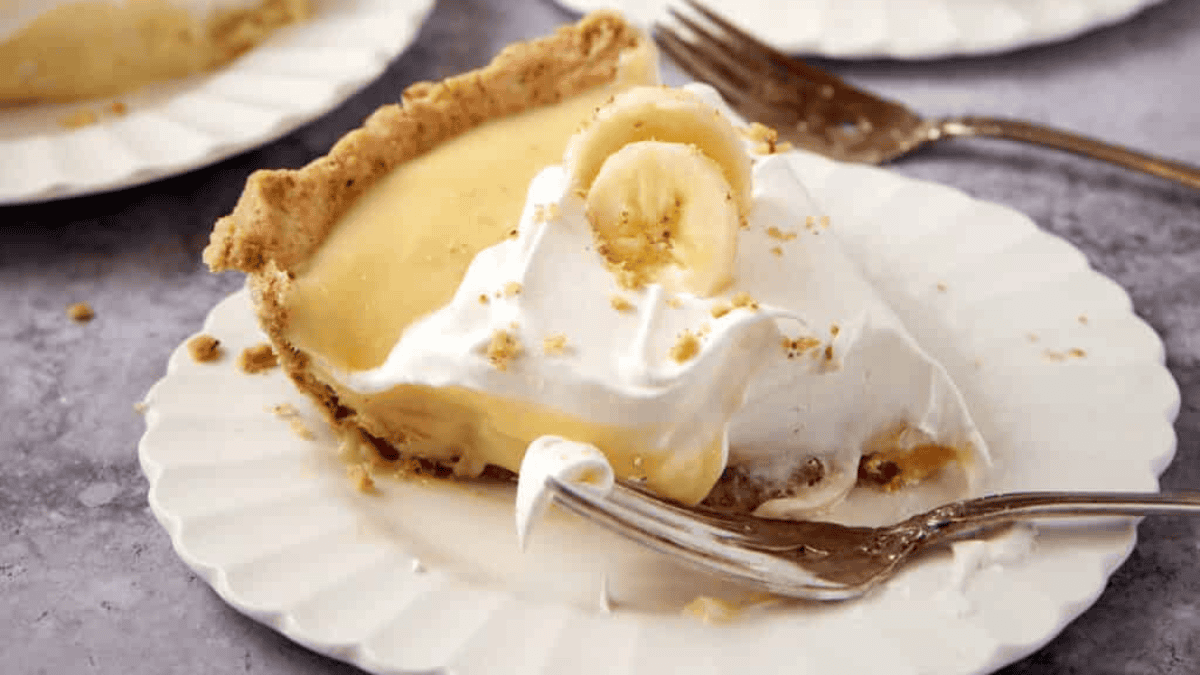 A slice of banana cream pie with a flaky crust, creamy filling, whipped topping, and banana slices on a white plate. A fork rests beside the pie, and another plate is visible in the background.