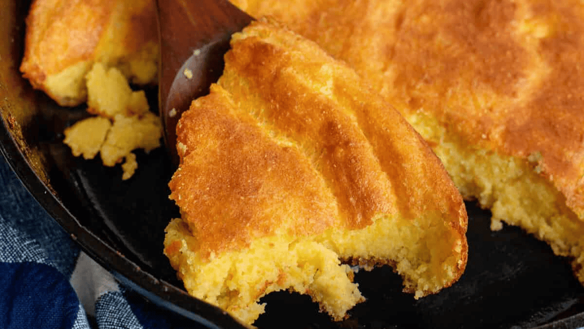 A slice of golden-brown cornbread being lifted from a cast iron skillet with a wooden spatula, showing its crumbly texture and moist interior.
