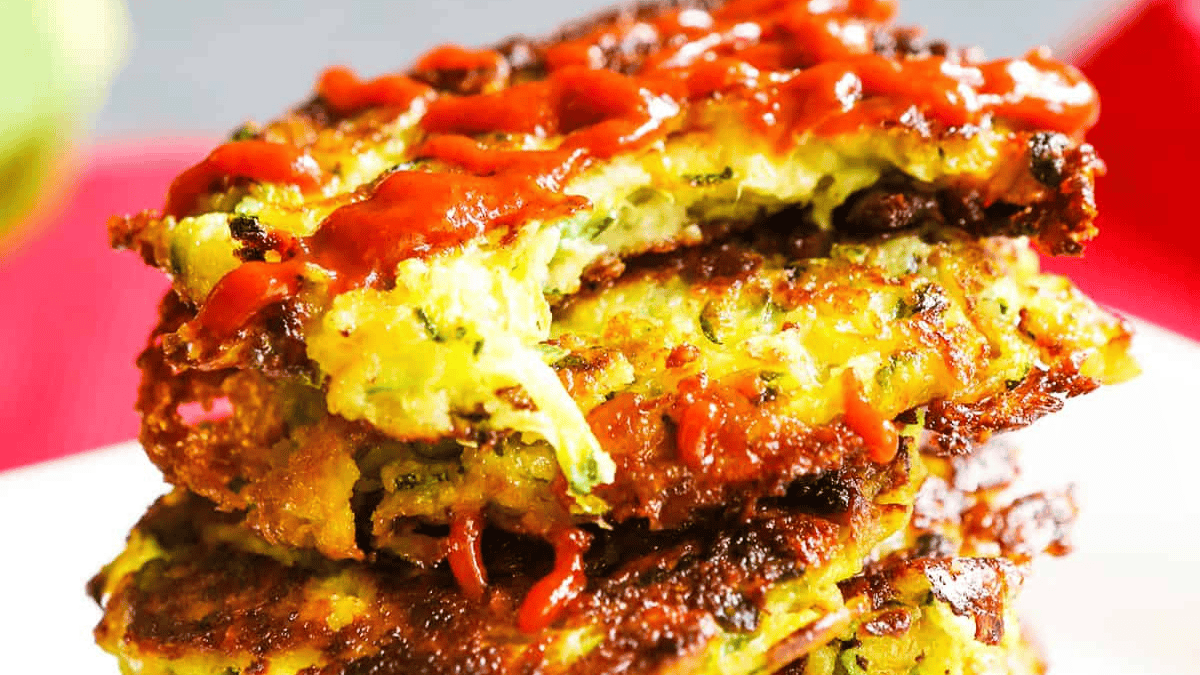 Three golden-brown zucchini fritters stacked on a plate, drizzled with bright red sauce. The fritters have a crispy texture and visible shreds of zucchini inside.