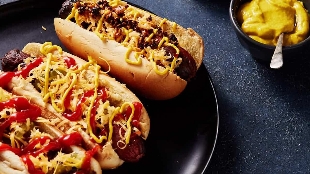 Two grilled hot dogs in buns topped with shredded cheese, ketchup, mustard, and chili, served on a black plate with a small bowl of yellow mustard and a spoon on the side.