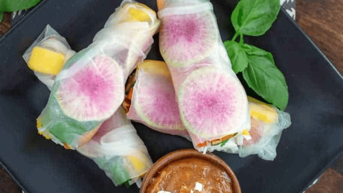 Three fresh spring rolls filled with colorful vegetables and slices of watermelon radish, served on a black plate with a small bowl of dipping sauce and fresh basil leaves.