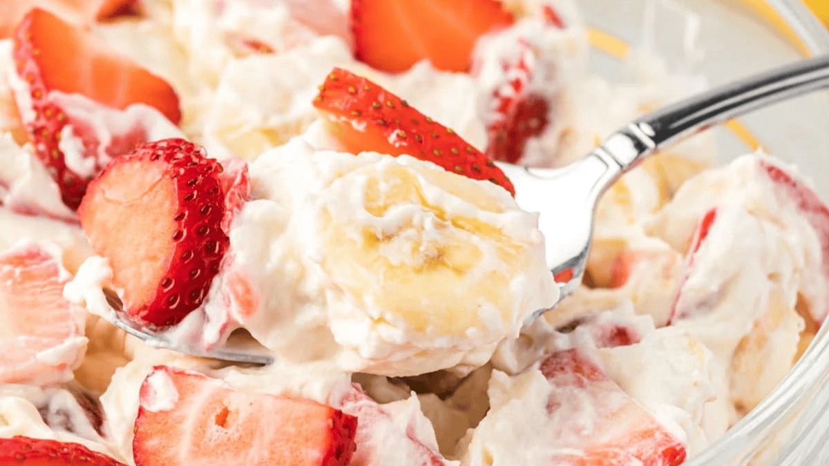 A close-up of a creamy fruit salad with sliced strawberries and bananas, served in a bowl with a spoon. The fruits are coated in a thick, white, creamy dressing.