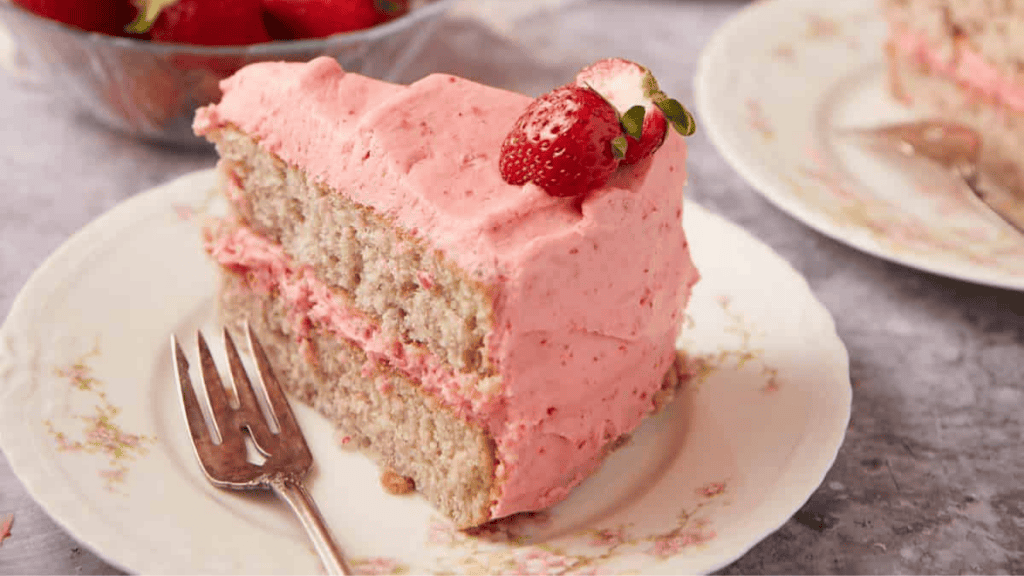 A slice of layered strawberry cake with pink frosting and a fresh strawberry on top sits on a floral plate with a fork. A bowl of strawberries and another plate with cake are in the background.