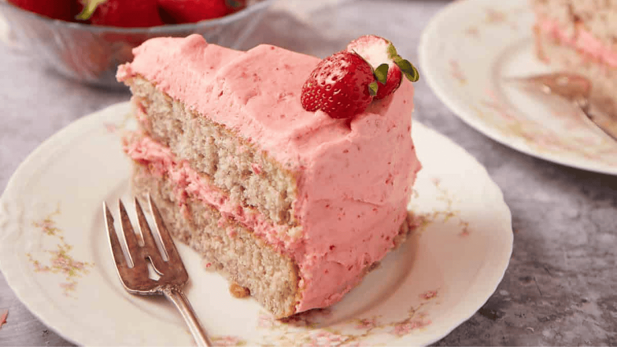 A slice of layered strawberry cake with pink frosting and a fresh strawberry on top sits on a floral plate with a fork. A bowl of strawberries and another plate with cake are in the background.