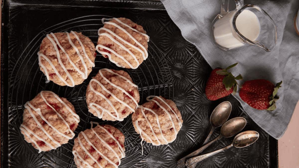 Six strawberry scones with white icing drizzle are arranged on a cooling rack. Nearby are vintage spoons, two strawberries, a pitcher of milk, and a gray cloth napkin on a dark textured surface.