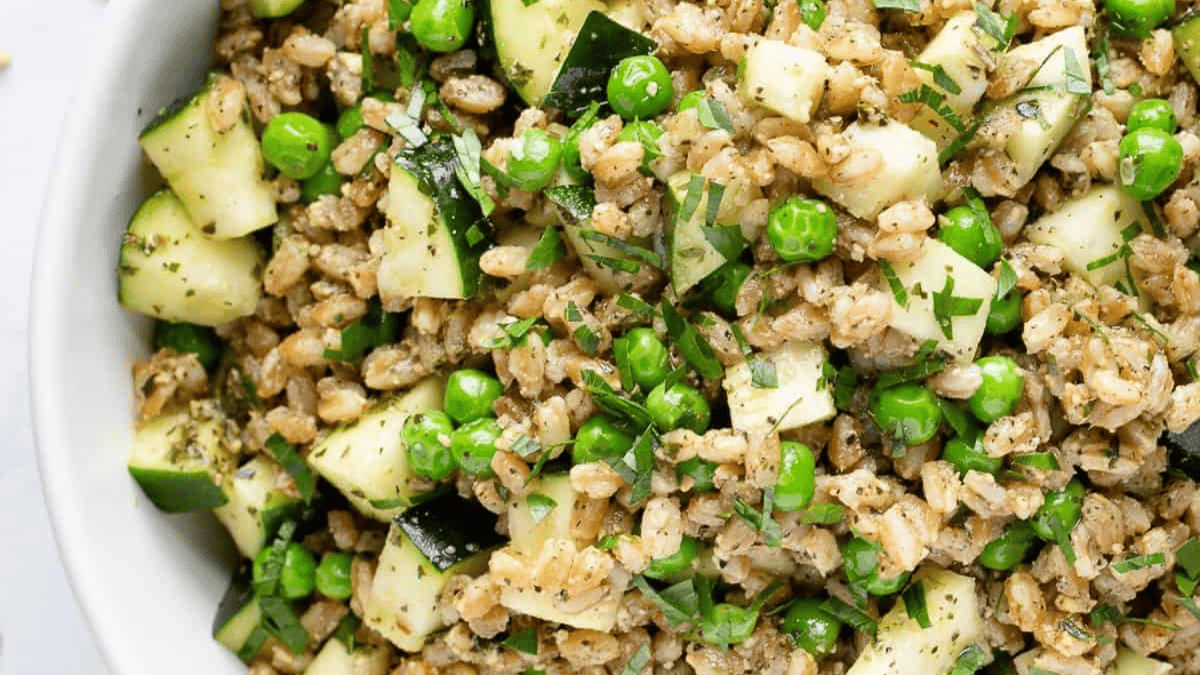 A close-up of a bowl filled with a salad made of barley, diced zucchini, green peas, and fresh herbs. The ingredients are mixed, showcasing a fresh and healthy grain salad.