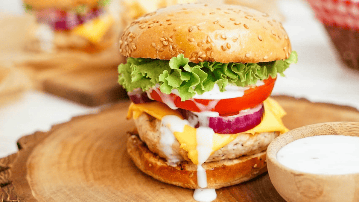A chicken burger with lettuce, tomato, red onion, cheddar cheese, and ranch dressing on a sesame seed bun, served on a wooden board with a small bowl of ranch dressing nearby.