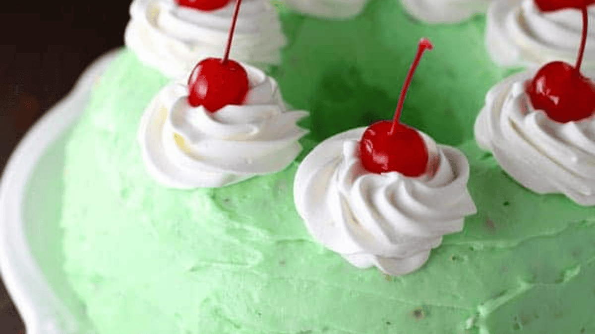 A close-up of a green frosted bundt cake topped with swirls of whipped cream and bright red maraschino cherries.