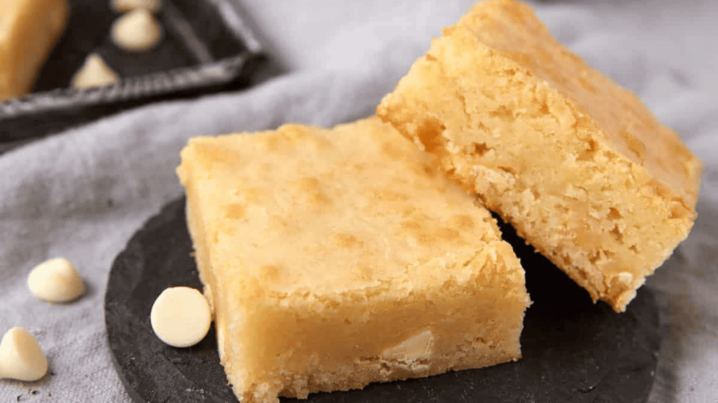 Two thick, square pieces of white chocolate blondies are stacked on a dark slate plate, with a few white chocolate chips scattered nearby. The blondies have a golden, slightly cracked top.