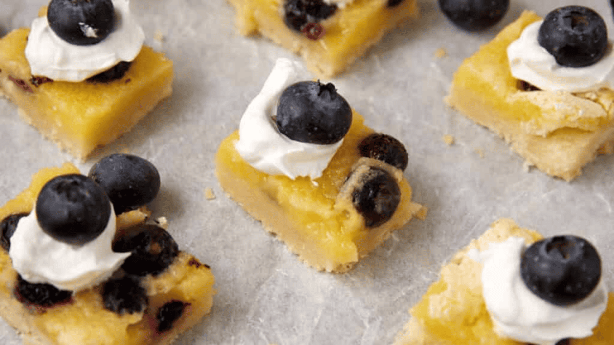 Squares of lemon bars topped with whipped cream and fresh blueberries are arranged on a sheet of parchment paper. The bars have a golden crust and a vibrant yellow filling.
