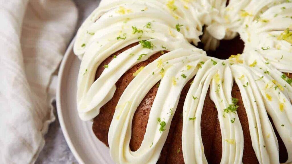 A close-up of a bundt cake topped with thick, white icing piped in loops and sprinkled with lemon and lime zest, sitting on a white plate next to a light-colored napkin.