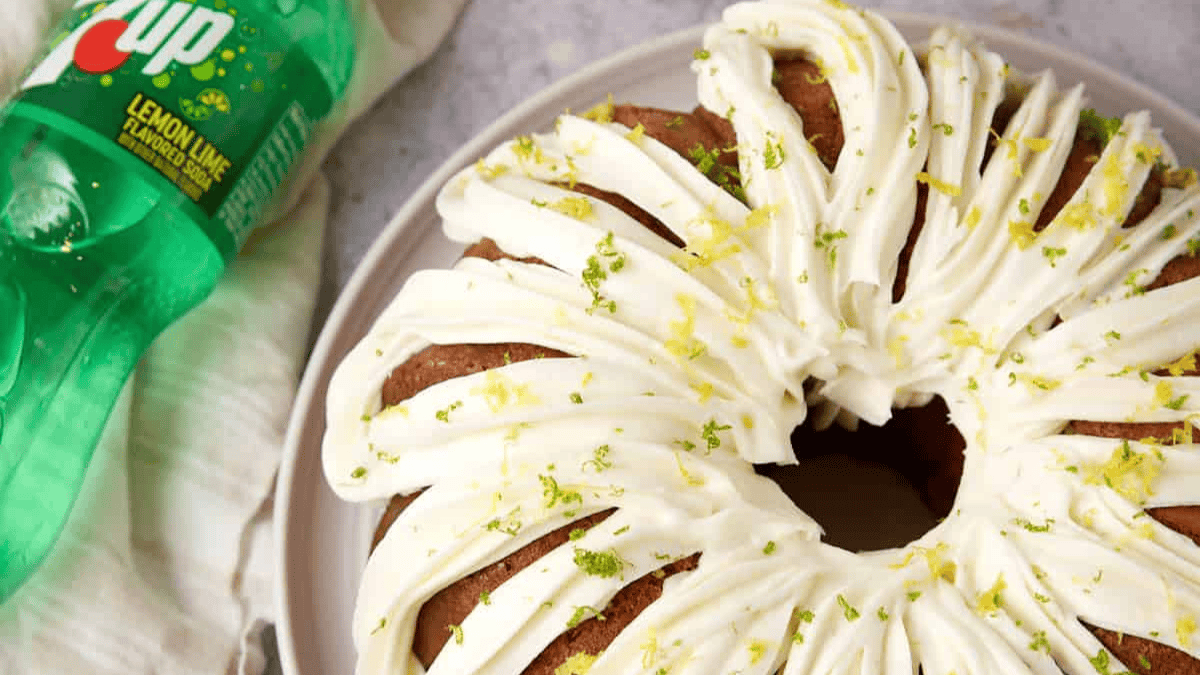 A bundt cake with white icing and lemon and lime zest on top sits on a plate next to a bottle of 7UP lemon-lime soda.