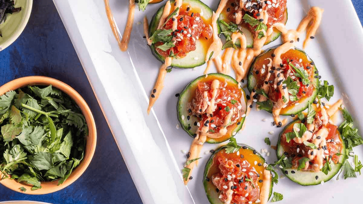 A rectangular white plate with cucumber slices topped with diced fish, sesame seeds, herbs, and drizzled sauce. A small bowl of fresh cilantro is nearby on the blue surface.