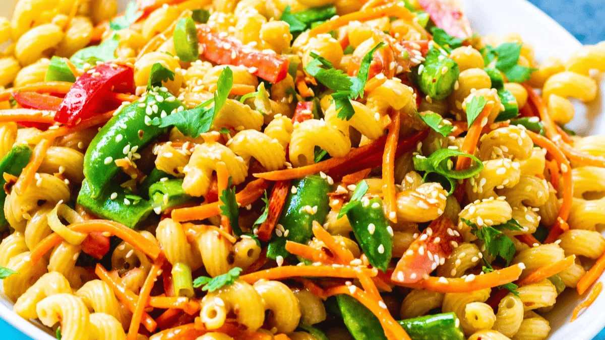 A close-up of colorful pasta salad with spiral noodles, sliced bell peppers, snap peas, shredded carrots, cilantro, and sesame seeds.