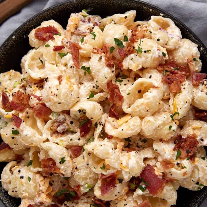 A black bowl filled with creamy macaroni salad topped with crispy bacon bits and chopped parsley, placed on a gray cloth next to a wooden spoon.