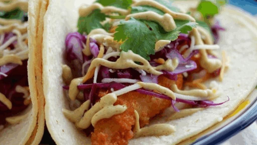 A close-up of a taco filled with crispy fried fish, shredded purple cabbage, fresh cilantro, and drizzled with a creamy sauce, all wrapped in a soft corn tortilla.