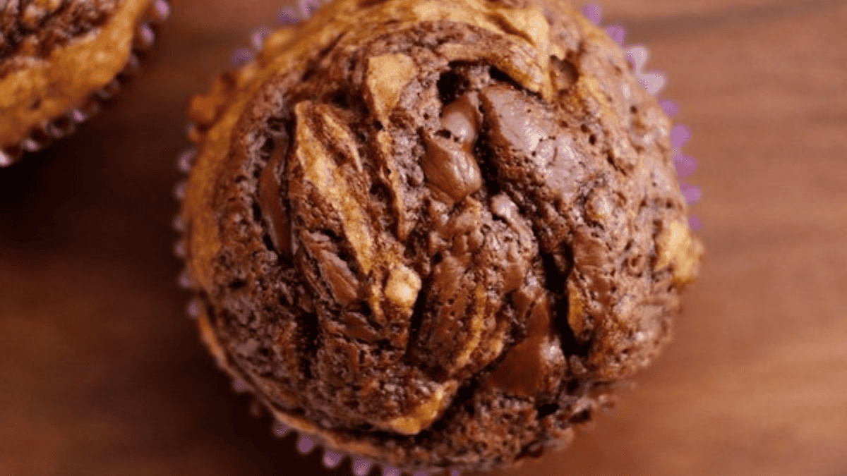 A close-up of a marbled muffin in a purple paper liner, featuring swirls of chocolate and light batter on a wooden surface.
