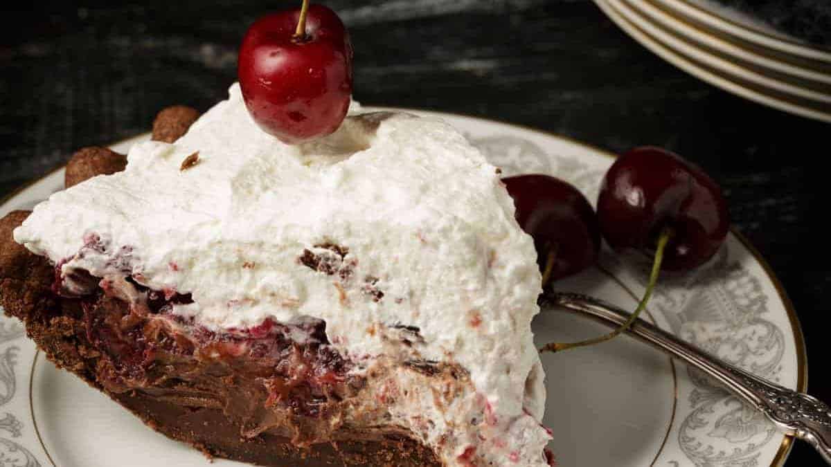 A slice of chocolate cherry pie topped with whipped cream and a cherry, served on a decorative plate with two fresh cherries and a fork beside it.