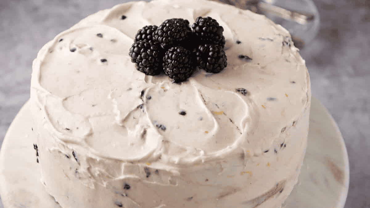 A round cake with creamy, light-colored frosting dotted with bits of fruit, topped with a small pile of fresh blackberries, displayed on a white plate.