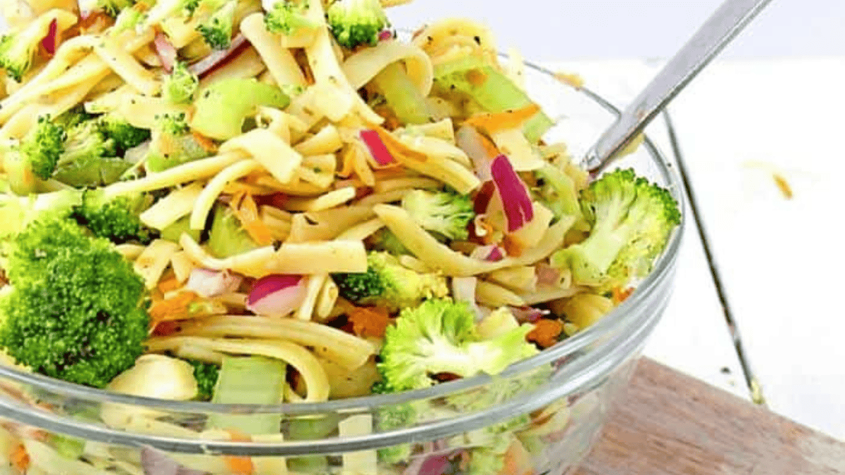 A glass bowl filled with pasta salad mixed with broccoli florets, red onion slices, carrots, and celery, with a serving spoon inside the bowl.