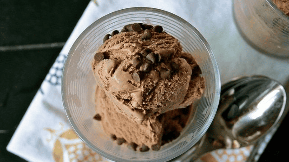 A glass bowl filled with scoops of chocolate ice cream topped with chocolate chips, placed on a white patterned cloth beside a metal spoon.