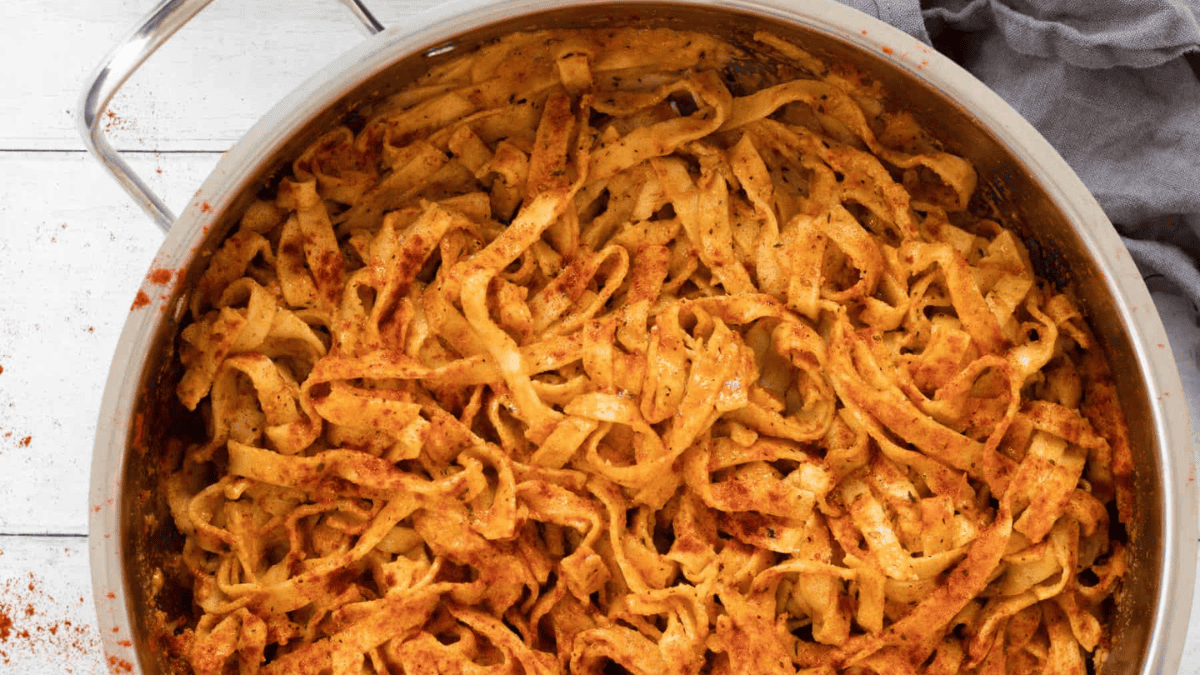 A large metal pan filled with creamy, orange-colored fettuccine pasta, coated in sauce and spices, sits on a light wooden surface next to a gray cloth.