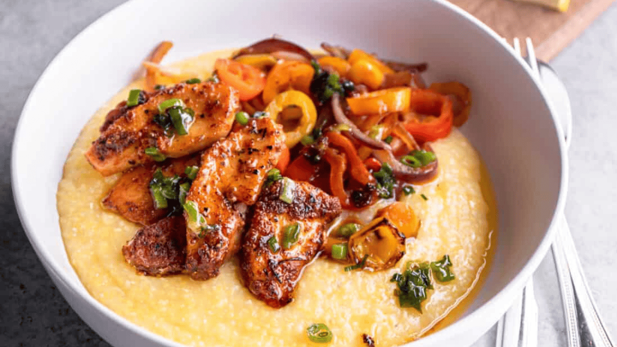 A bowl of creamy yellow grits topped with seared pieces of seasoned chicken, sautéed colorful bell peppers, red onions, and garnished with chopped green onions. A fork and knife are placed beside the bowl.