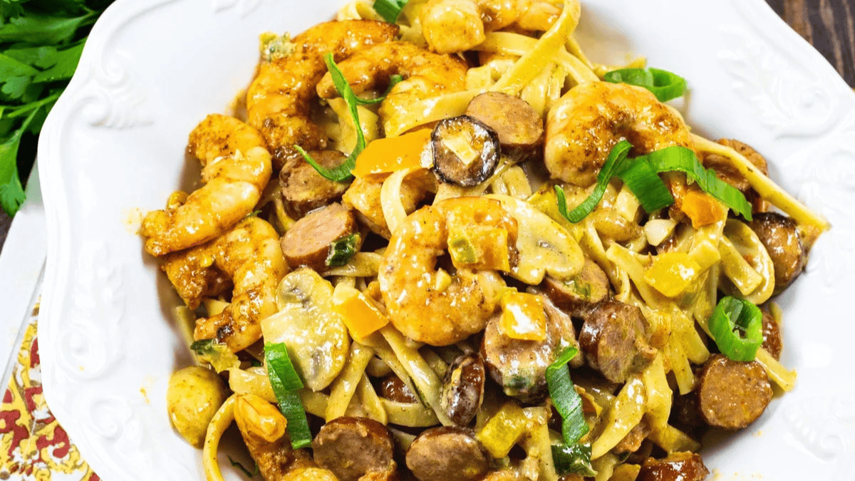 A white bowl filled with creamy pasta topped with shrimp, sliced sausage, sautéed vegetables, and garnished with chopped green onions.