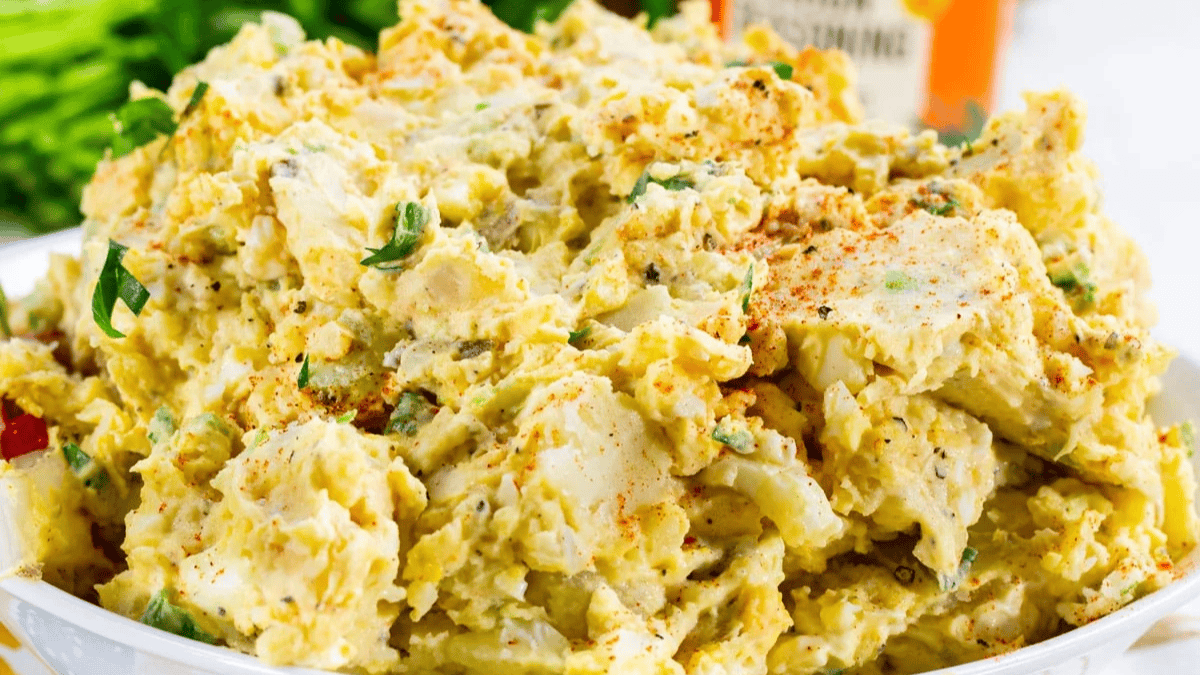 A close-up of a bowl filled with creamy potato salad, garnished with chopped green onions and a sprinkle of paprika, with some leafy greens and a container blurred in the background.