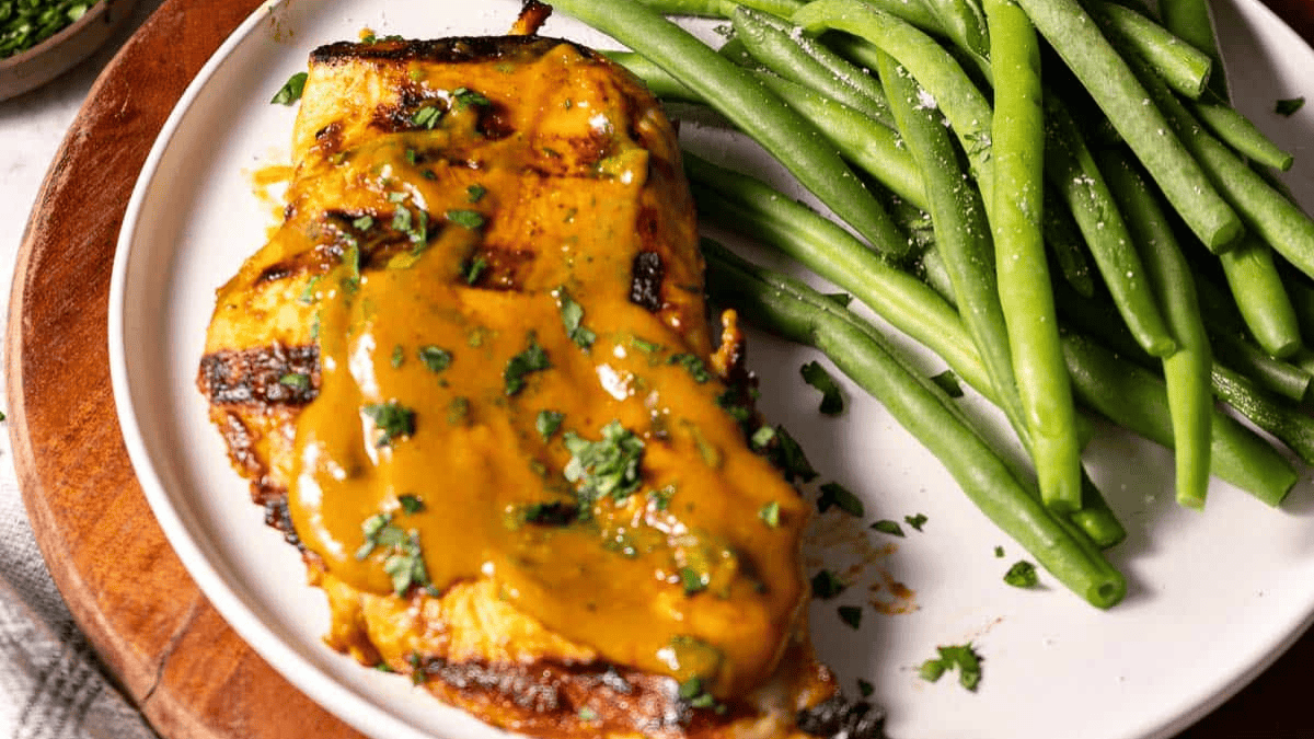 A plate with grilled chicken breast topped with a yellowish sauce and chopped herbs, served alongside cooked green beans.