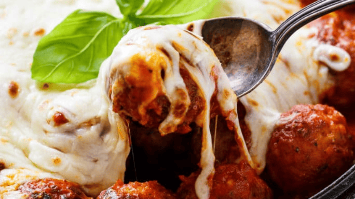 A close-up of cheesy baked meatballs in marinara sauce, topped with melted mozzarella cheese and garnished with a fresh basil leaf. A spoon is lifting a meatball, stretching melted cheese.
