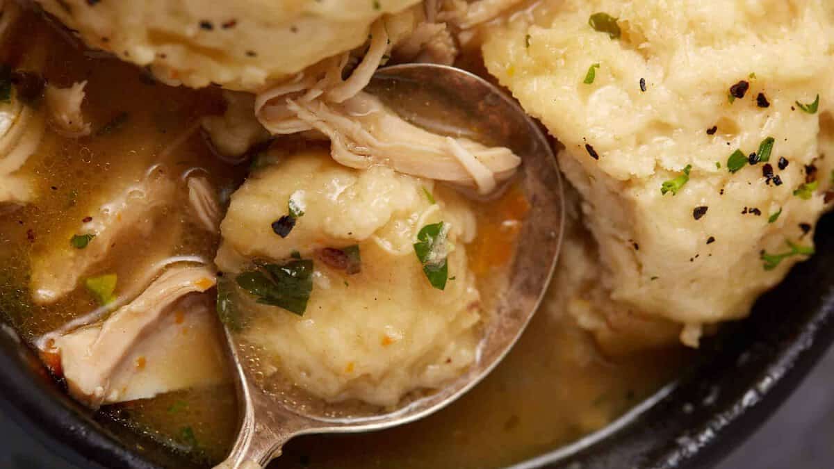 Close-up of a bowl of chicken and dumplings. A spoon rests in the bowl, lifting one of the dumplings. The golden broth, shredded chicken, and sprinkled herbs on top are visible, giving a savory and comforting appearance to the dish.