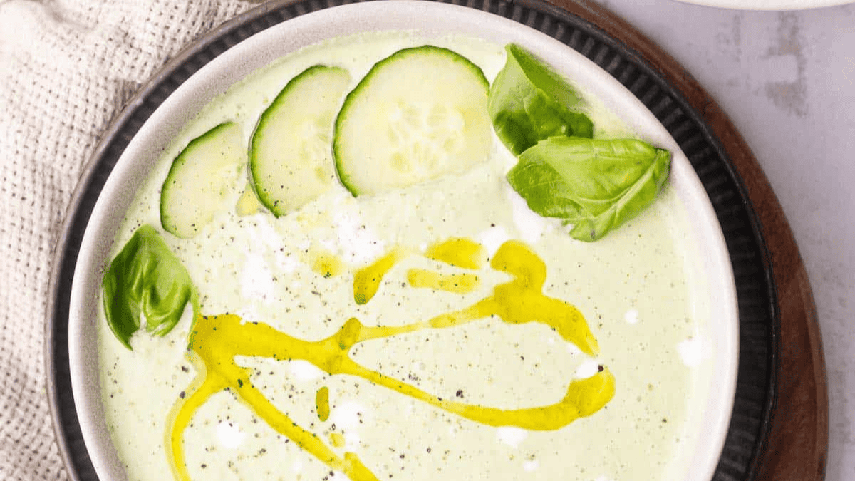 A bowl of creamy green soup topped with cucumber slices, fresh basil leaves, a drizzle of olive oil, and a sprinkle of black pepper. The bowl is set on a dark plate with a textured cloth beside it.