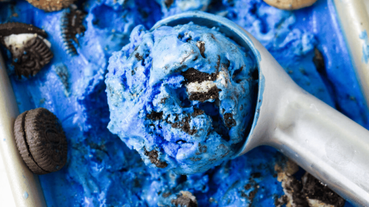 A metal ice cream scoop holds bright blue cookies and cream ice cream with chunks of chocolate sandwich cookies, surrounded by more ice cream and broken cookies in a metal tray.