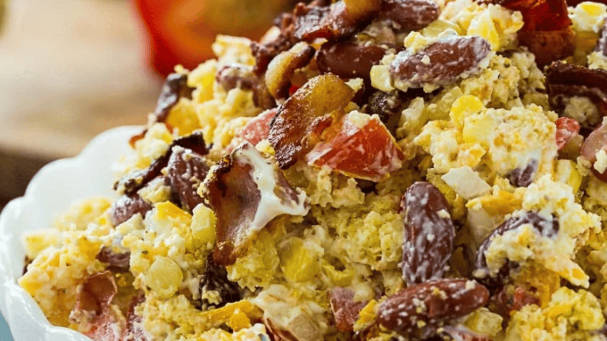 A close-up of a creamy salad with kidney beans, corn, diced tomatoes, chopped onions, shredded cheese, and crispy bacon pieces in a white bowl.